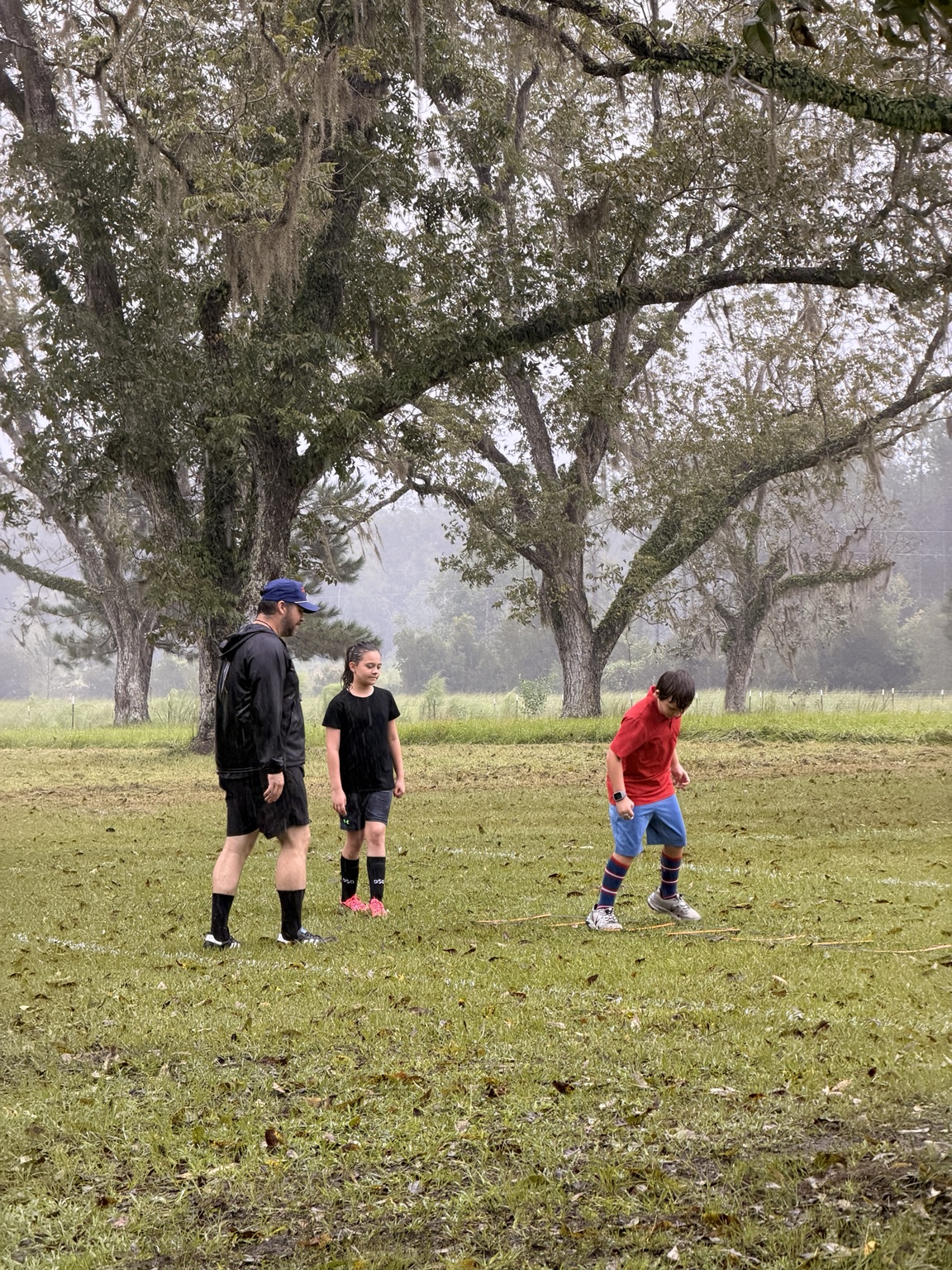 An ORVIT coach guiding two students through soccer training in the rain beneath the pecan trees